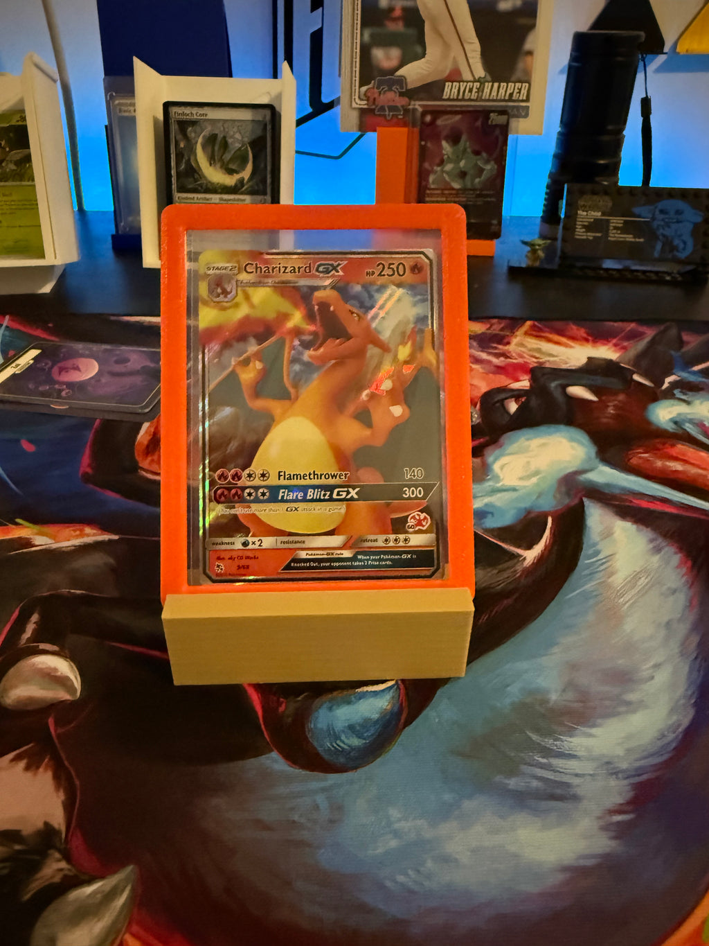 Trading card in a holder on a colorful card game mat with other cards and game components in the background.